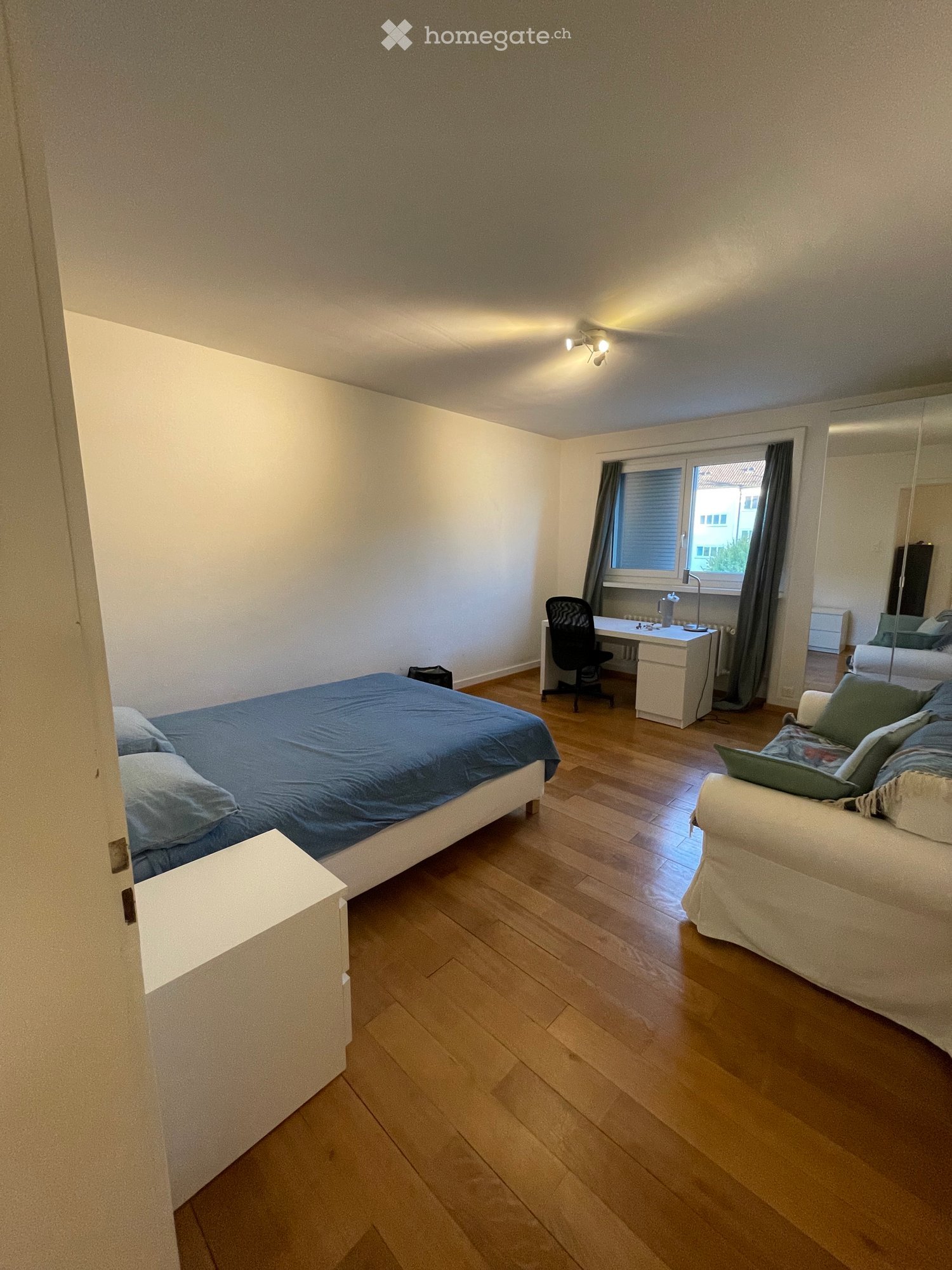 Well-lit bedroom with wooden flooring, a bed with blue bedding, a sofa with green cushions, a desk with a chair, and a large mirror on the wall.
