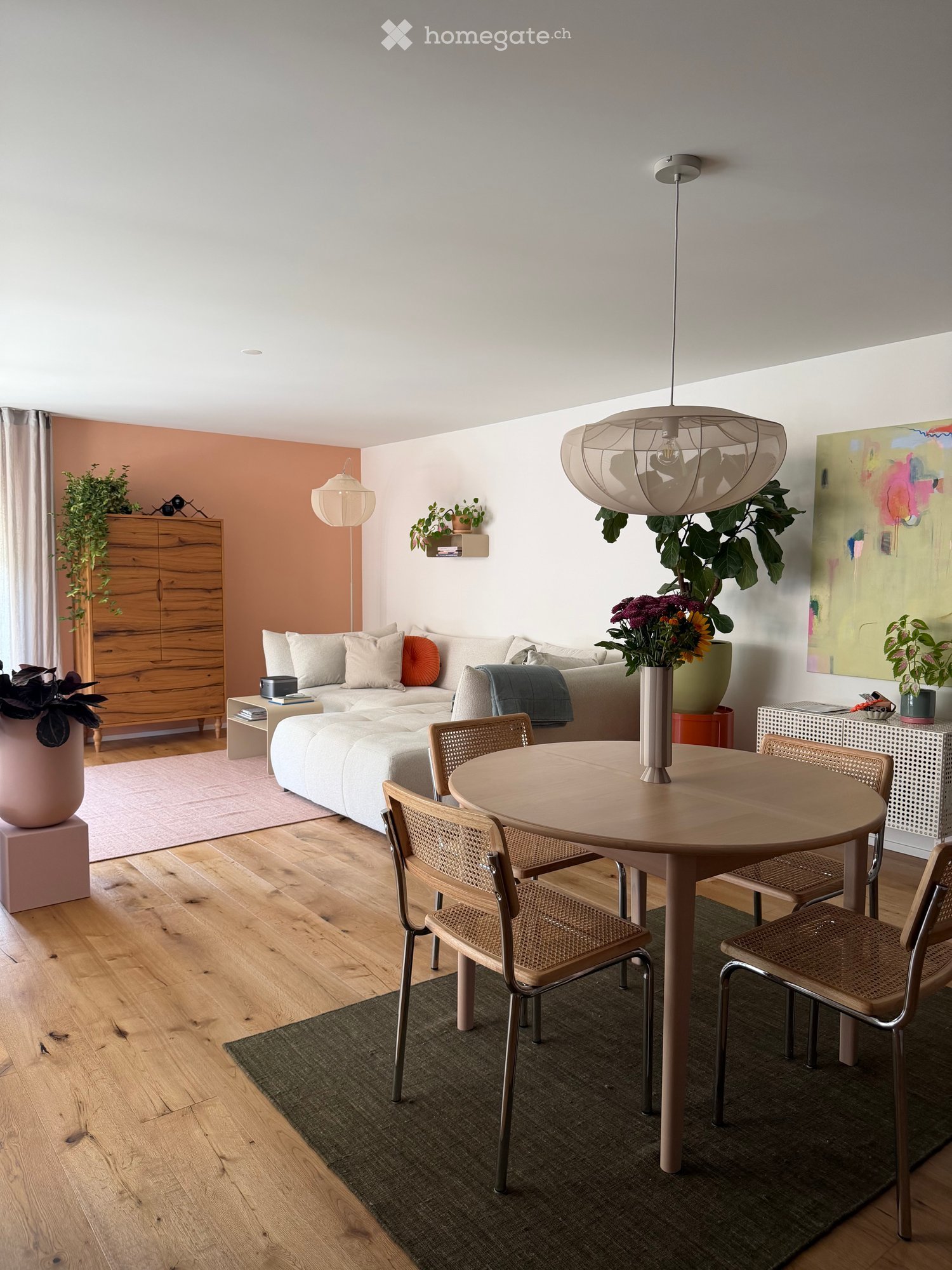 Modern living room with a white sofa, round wooden dining table, wicker chairs, pendant light, and decorative plants.