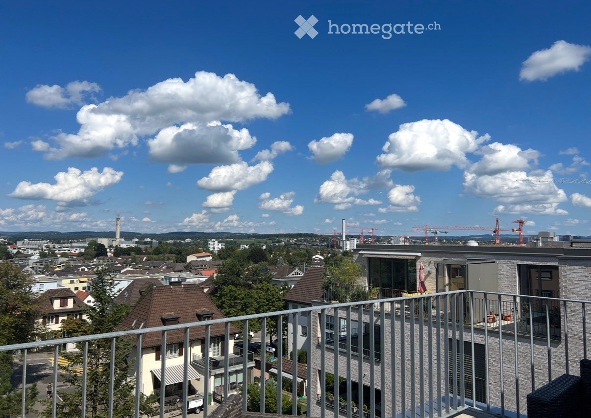 Balcony overlooking a cityscape with residential buildings, trees, and distant construction cranes under a partly cloudy sky.