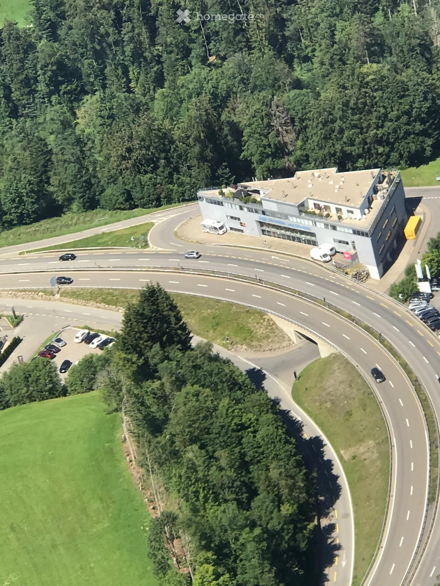 Aerial view of a modern, multi-story building with a flat roof, surrounded by a parking lot and a winding road. The building has large windows and a sign on the front. The surrounding area includes trees, a grassy field, and a road with several cars.