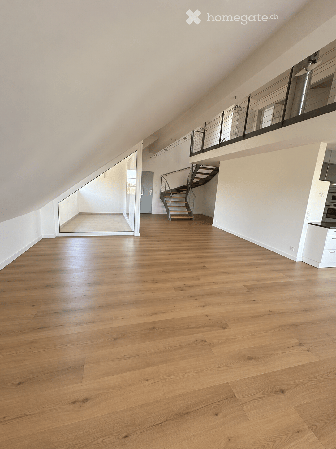 An empty, unfurnished room with light wooden flooring, white walls, and a staircase leading to an upper level. The space features a modern design with large windows and a glass door leading to another area.