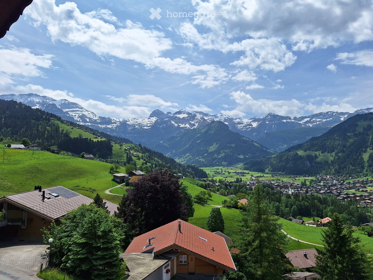 Panoramic view of a village nestled in a lush green valley surrounded by snow-capped mountains. The foreground shows a house with a red-tiled roof and a driveway, while the background features rolling hills, scattered houses, and a winding road.