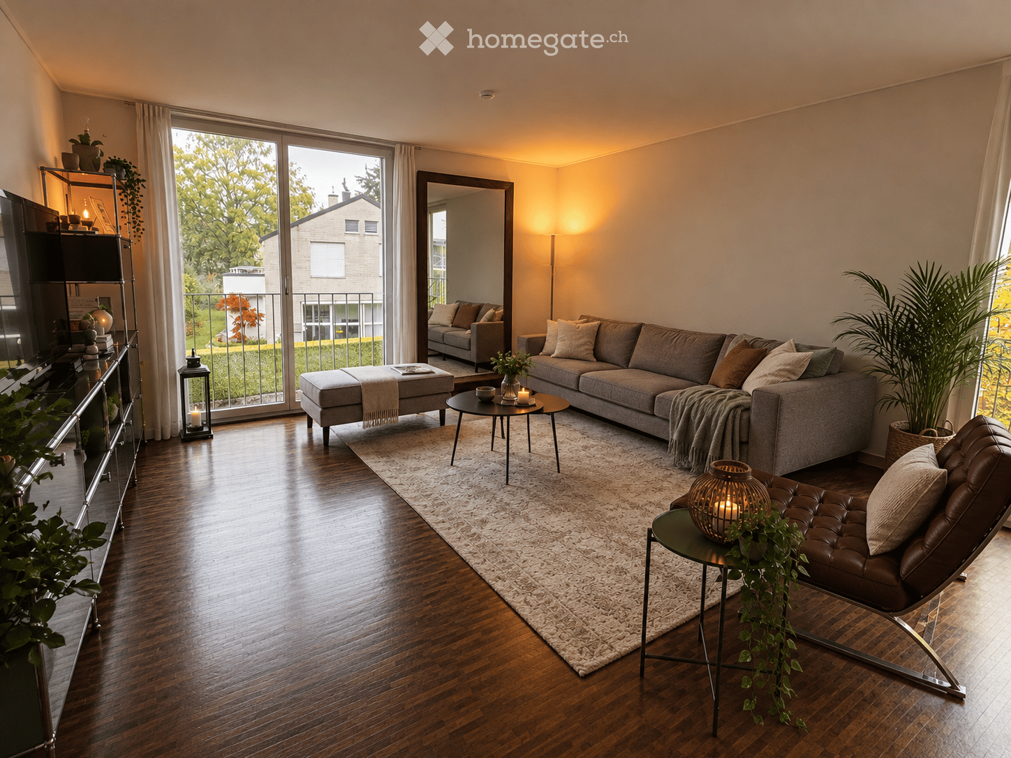 Modern living room with hardwood floors, large windows, grey sectional sofa, ottoman, coffee table, leather chair, floor lamp, and decorative plants.