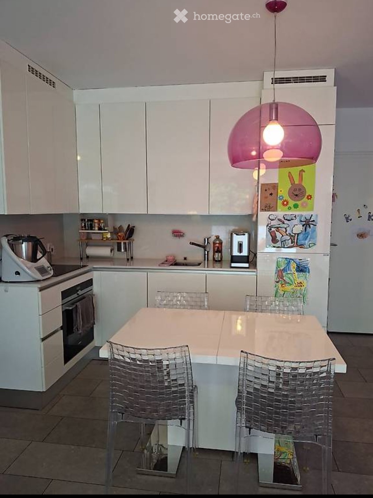 Modern kitchen with white cabinets, a central island table with four transparent chairs, a refrigerator with children's drawings, and various appliances including a blender and oven.