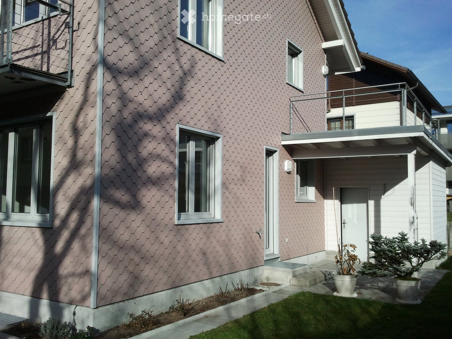 Two-story house with pink siding, white-framed windows, and a balcony on the upper level. The entrance has a small porch with steps leading up to a white door. Potted plants are placed near the entrance.