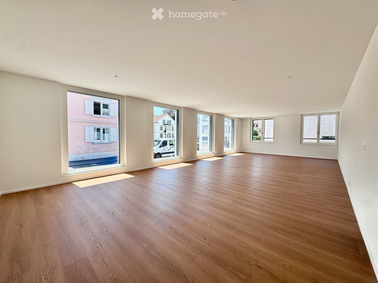 Empty room with wooden flooring and large windows, showing natural light and a view of the street.