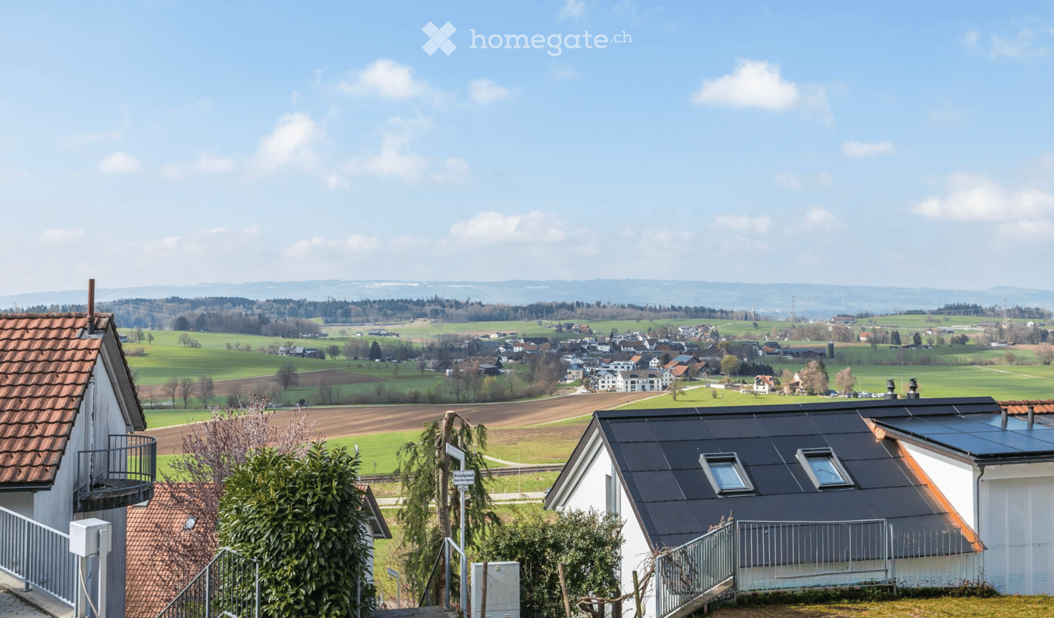 A peaceful rural village scene with houses, green fields, and distant hills under a clear blue sky.