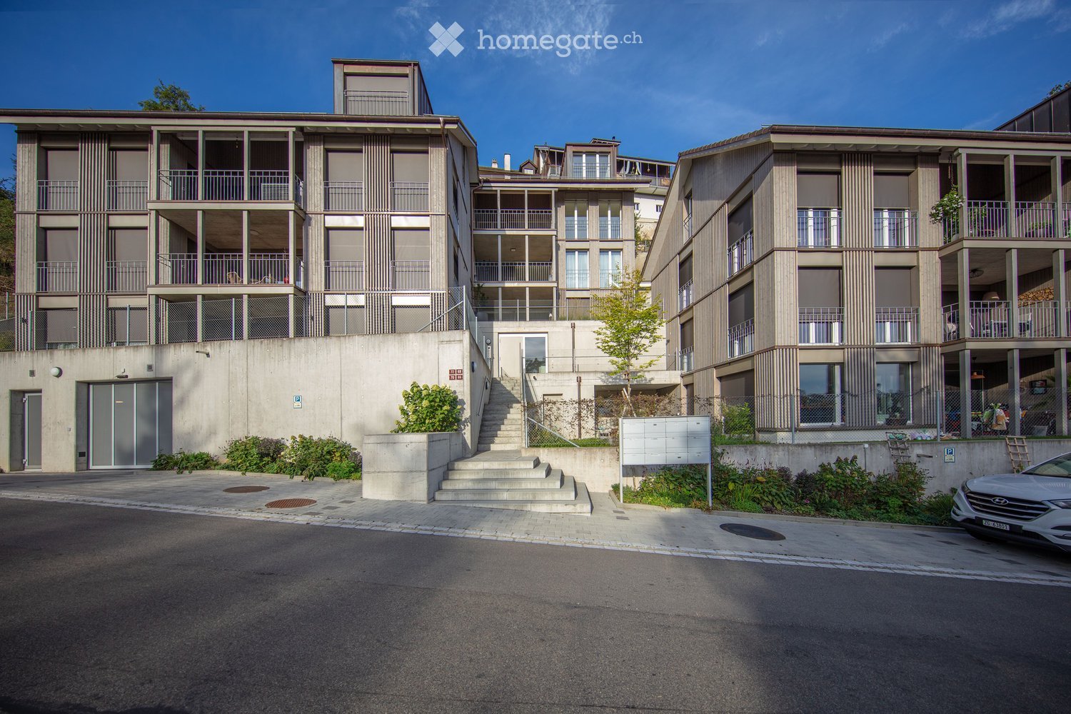 Modern multi-story residential building with balconies, concrete facade, and a raised ground floor entrance.