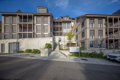 Modern multi-story apartment building with balconies, concrete facade, and a central staircase leading to the entrance.