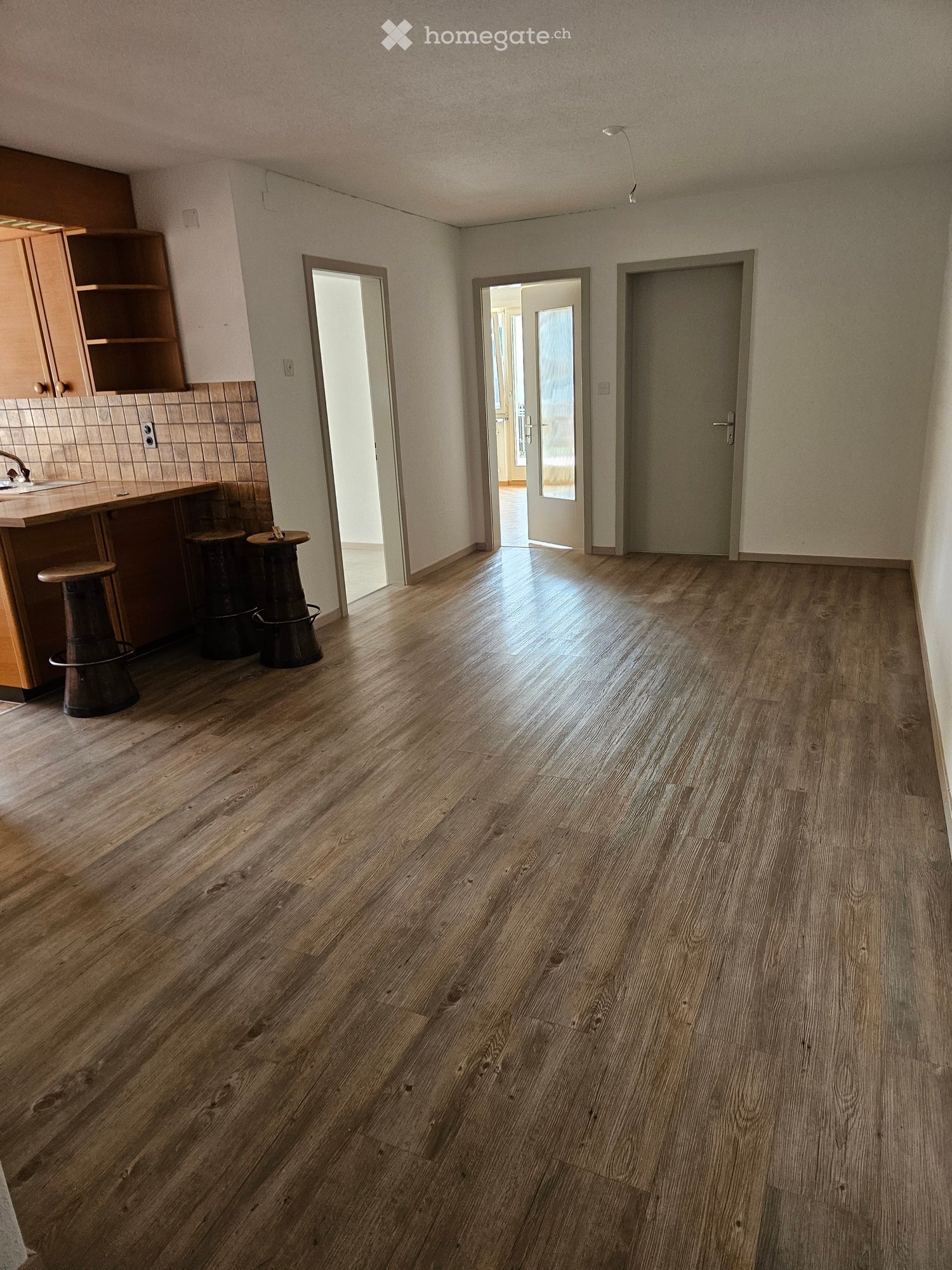 Empty room with wooden flooring, light-colored walls, and several doors leading to other spaces. There is a small kitchen area with cabinets and a sink visible on the left side.