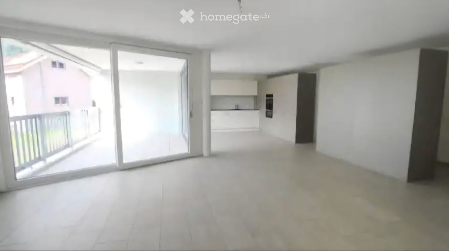 Empty living space with white walls, large sliding glass doors leading to a balcony, and a view of a neighboring building.