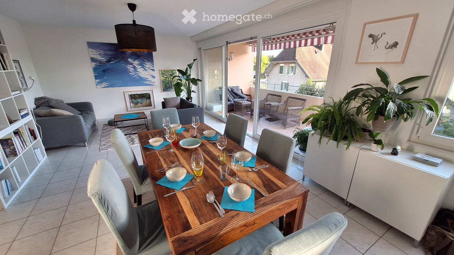 Bright living and dining area with a grey sofa, wooden dining table, chairs, wine glasses, and decorative plants. Large sliding glass doors open to a balcony with outdoor seating.