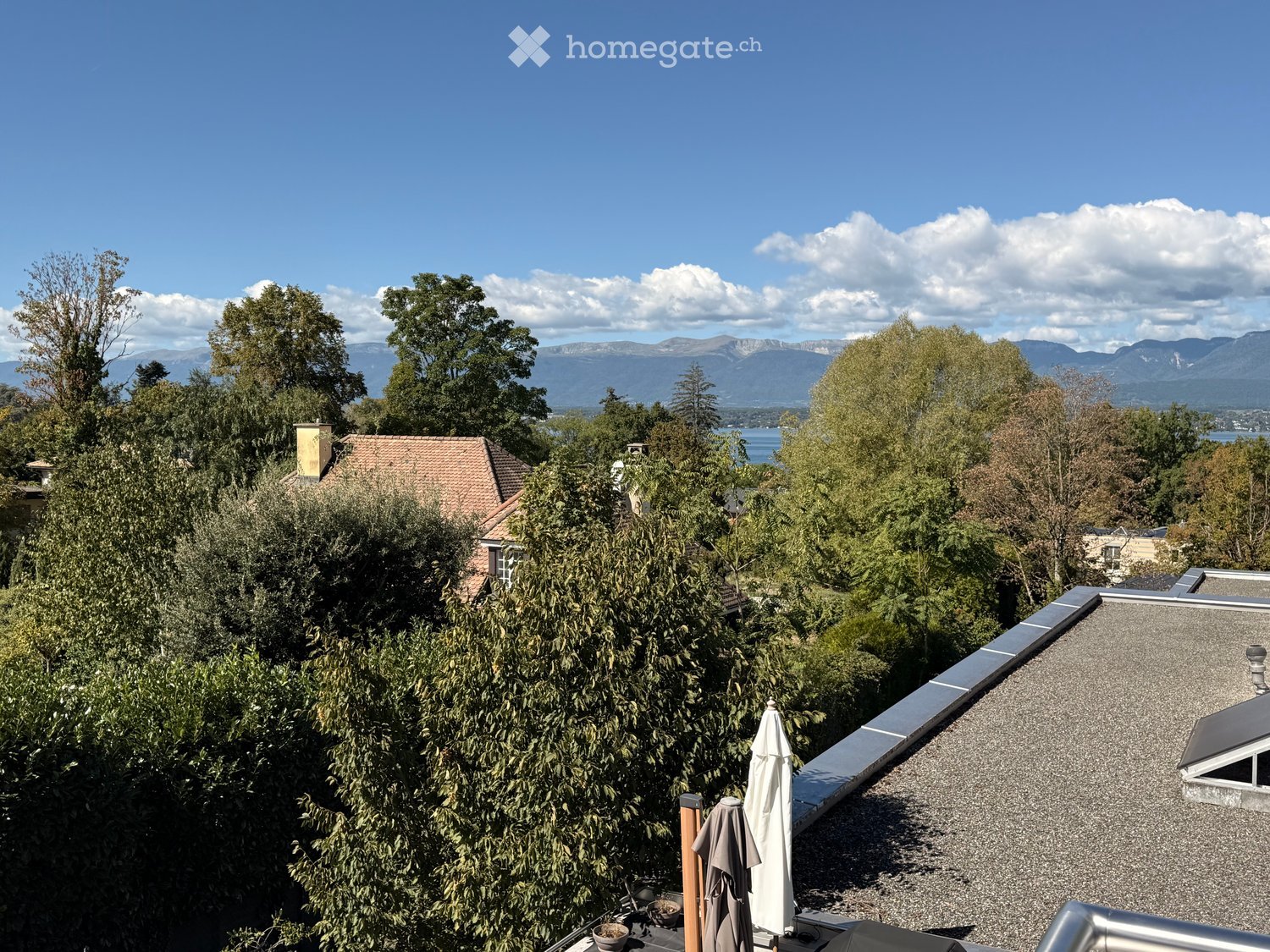 A rooftop terrace with a view of a lake and mountains, surrounded by trees and a house with a red-tiled roof.