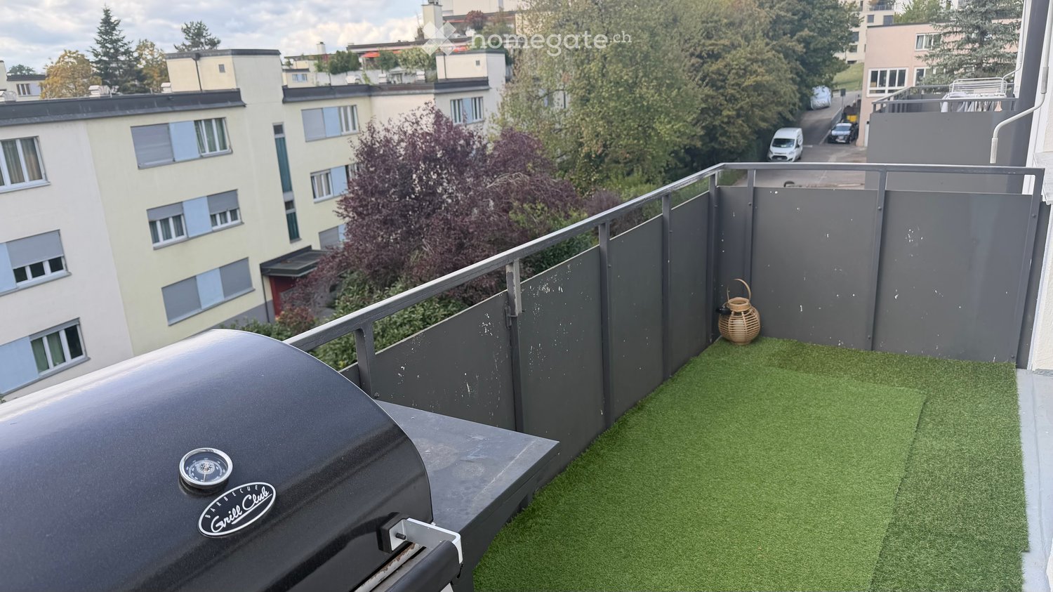 Balcony with artificial grass, a grill, and a small woven basket. The railing is modern and metallic, and the view shows neighboring buildings and trees.