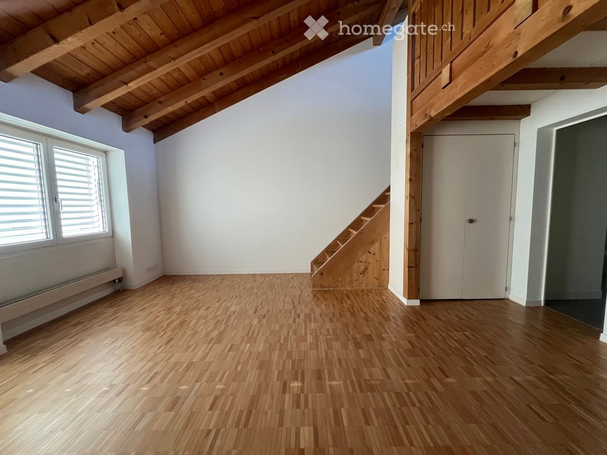 Empty room with wooden flooring, white walls, exposed wooden ceiling beams, a staircase leading upwards, and a closed door.