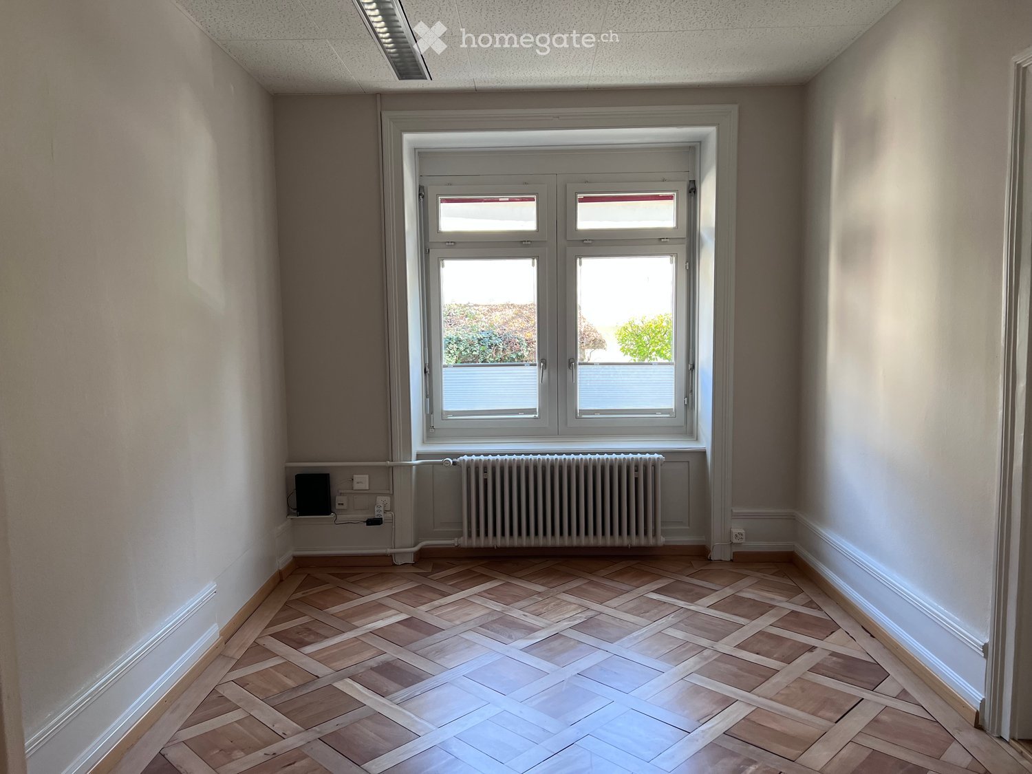 Empty room with wooden herringbone flooring, white walls, large window with double glazing, radiator, and electrical outlets.