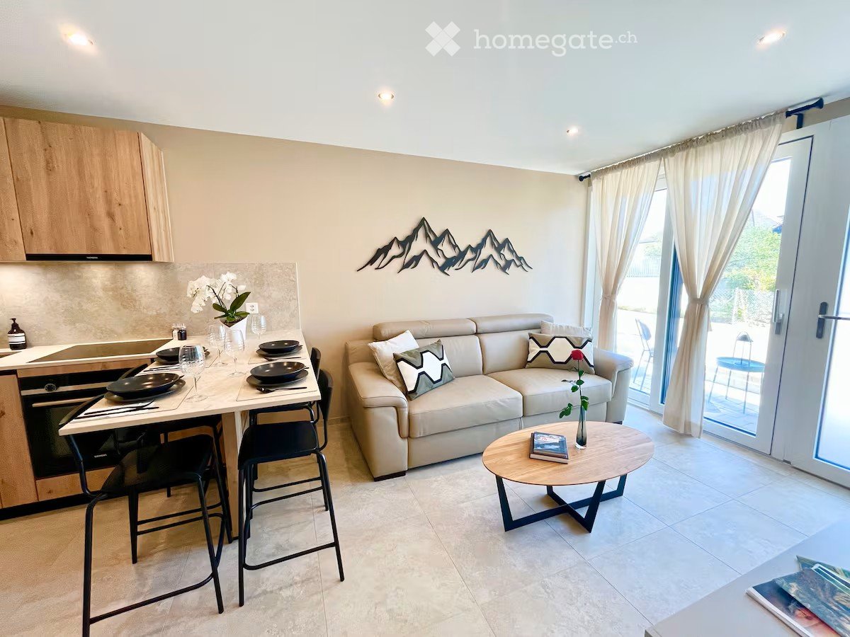 Furnished Room. Modern open-plan living and dining area with a cream sofa, coffee table, dining table with stools, kitchen area with cabinets and appliances, and decorative wall art.