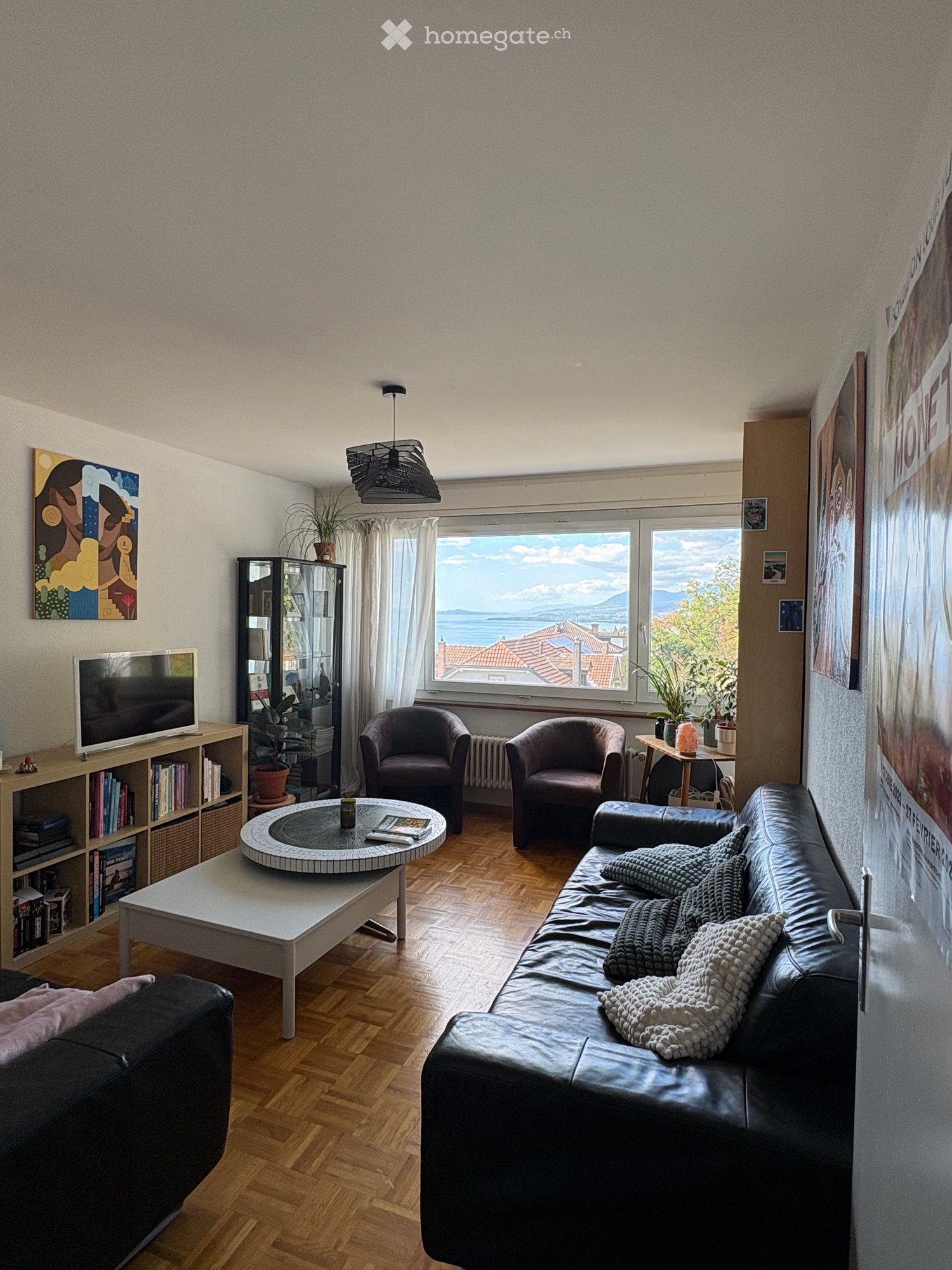 Modern living room with wooden flooring, large windows offering a scenic view, a black leather sofa with blankets, a white coffee table, two armchairs, a bookshelf, and a television.