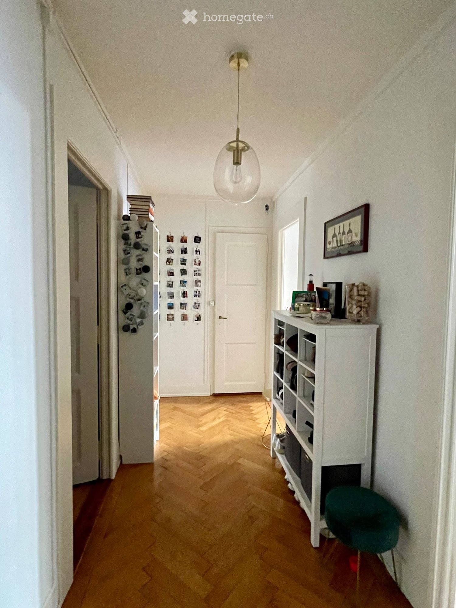 A narrow hallway with wooden herringbone flooring, white walls, and a modern pendant light fixture. A white shelving unit with books and decorative items is visible on the right, along with a green chair and a framed picture on the wall.