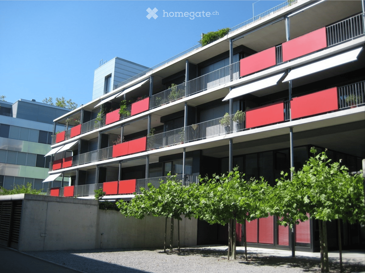 Modern multi-story residential building with red balconies, metal railings, and greenery in front.