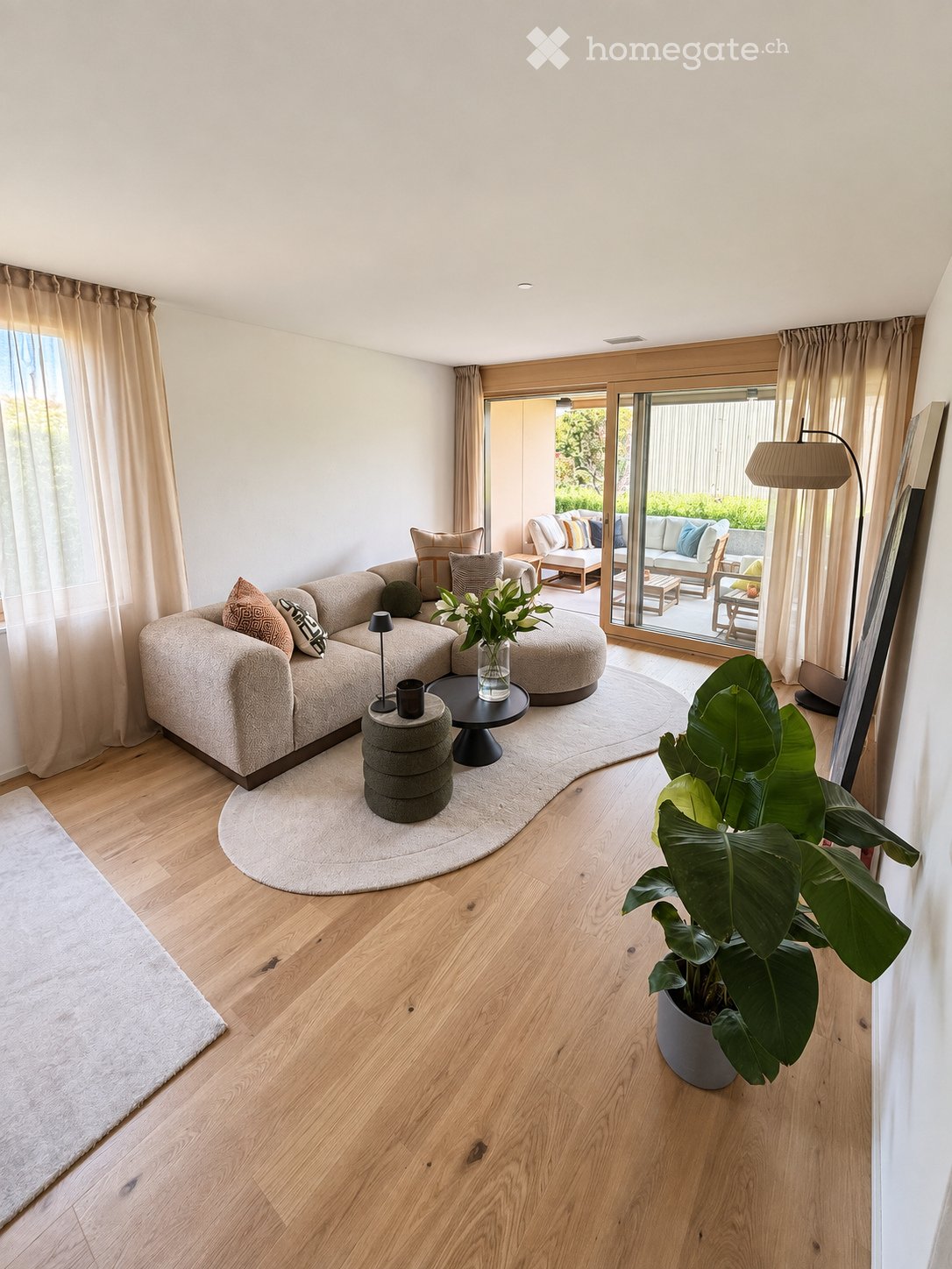 Modern living room with light wooden flooring, large sectional sofa, coffee table, floor lamp, and a large potted plant near the sliding glass doors leading to an outdoor area.