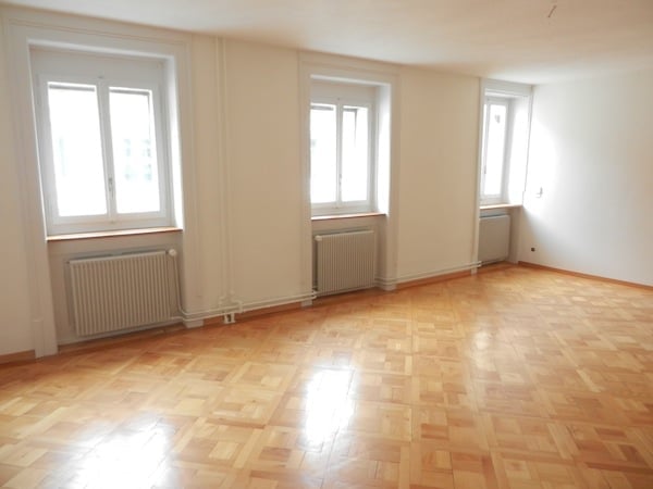 Empty room with wooden parquet flooring, white walls, and three windows. Radiators are visible under each window.