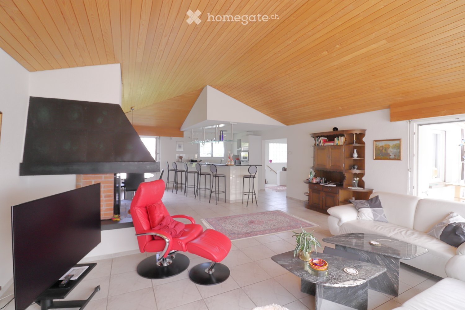 Modern living room with a white sofa, red leather chair, glass coffee table, TV screen, and a small kitchen area with bar stools.