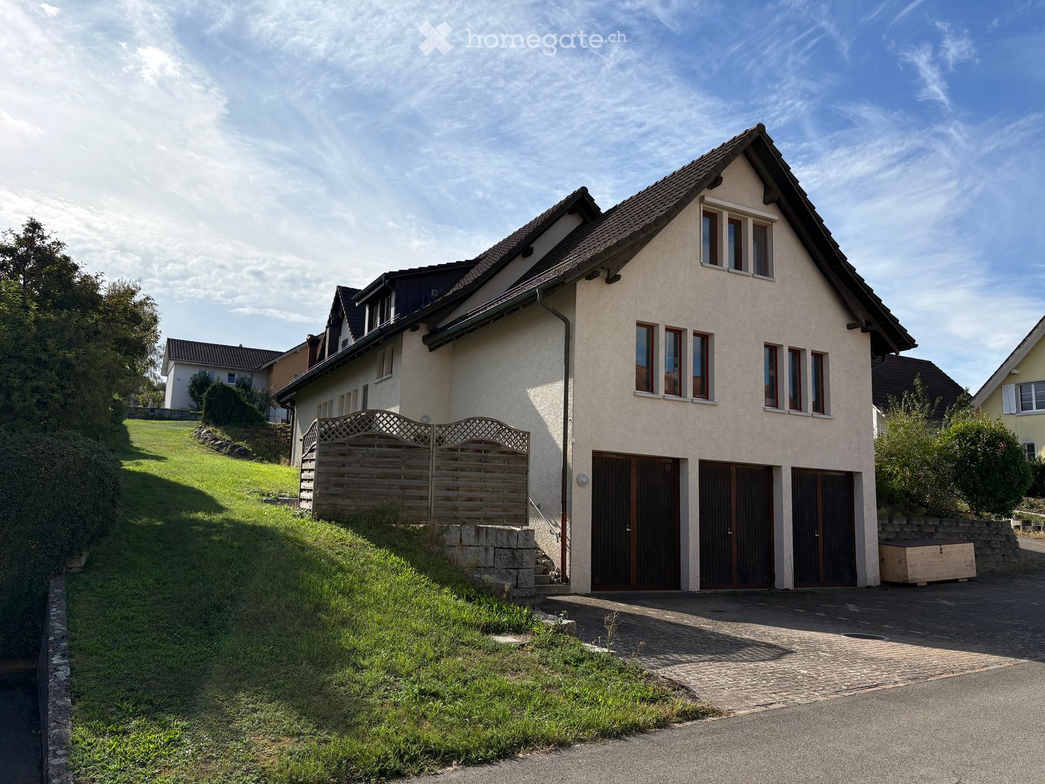 Two-story house with beige facade, dark brown garage doors, and multiple windows. The house is surrounded by a wooden fence and greenery, with a paved driveway leading to the garage.