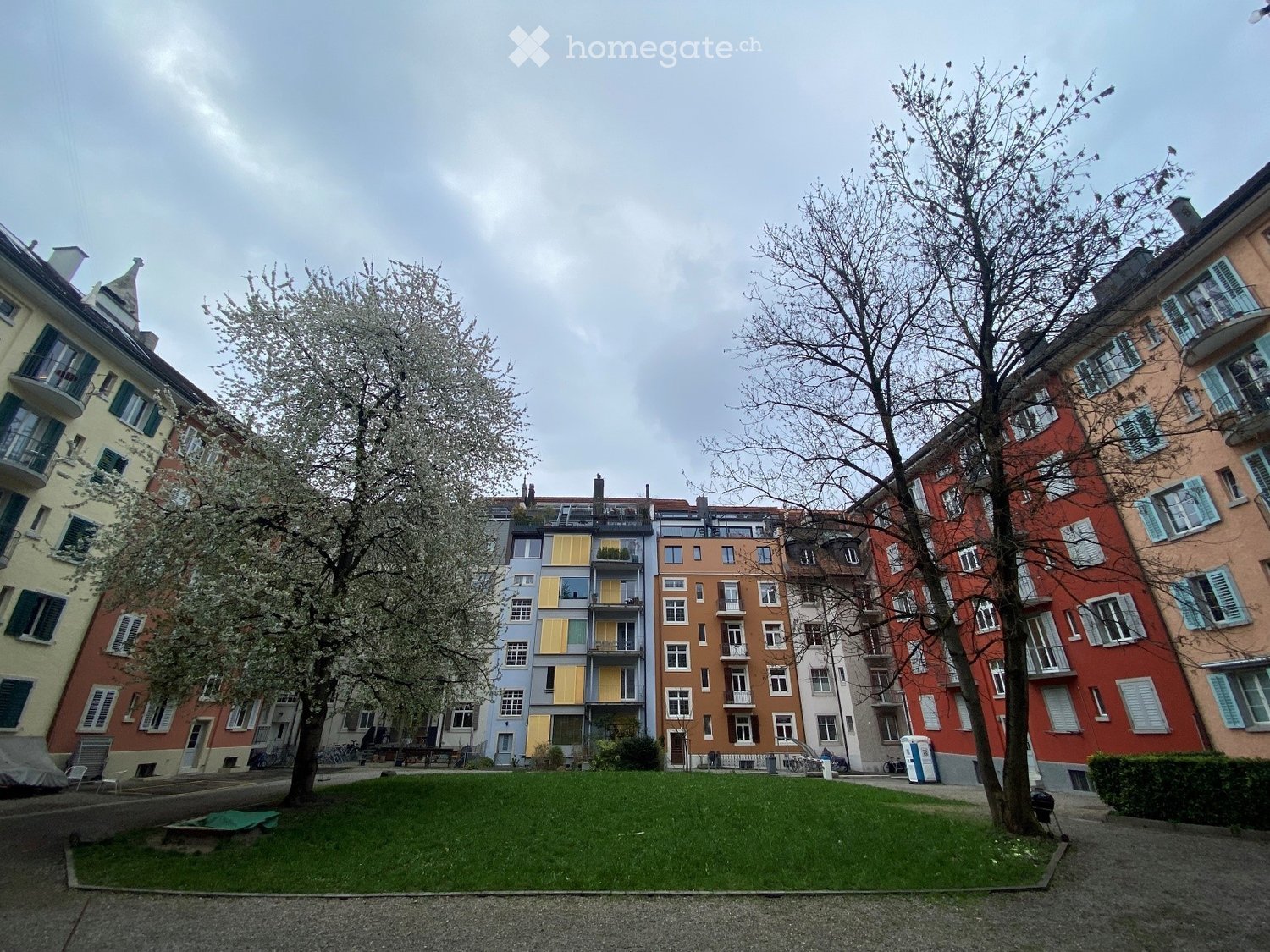 A park area surrounded by colorful residential buildings with balconies and shutters, featuring trees and a grassy lawn.