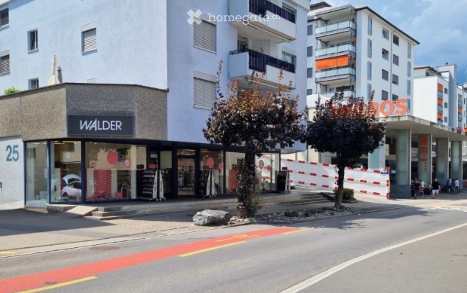 Modern commercial building with storefronts, including a 'WALDER' store and a 'ROS' store, surrounded by trees and pedestrians.