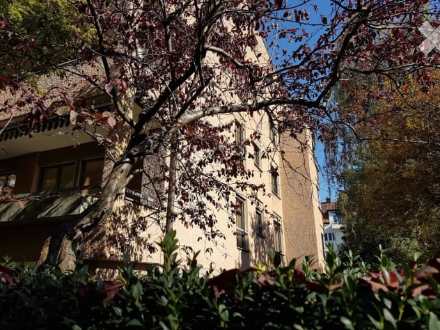 A multi-story residential building with a beige facade, multiple windows, and a tree partially obscuring the view.