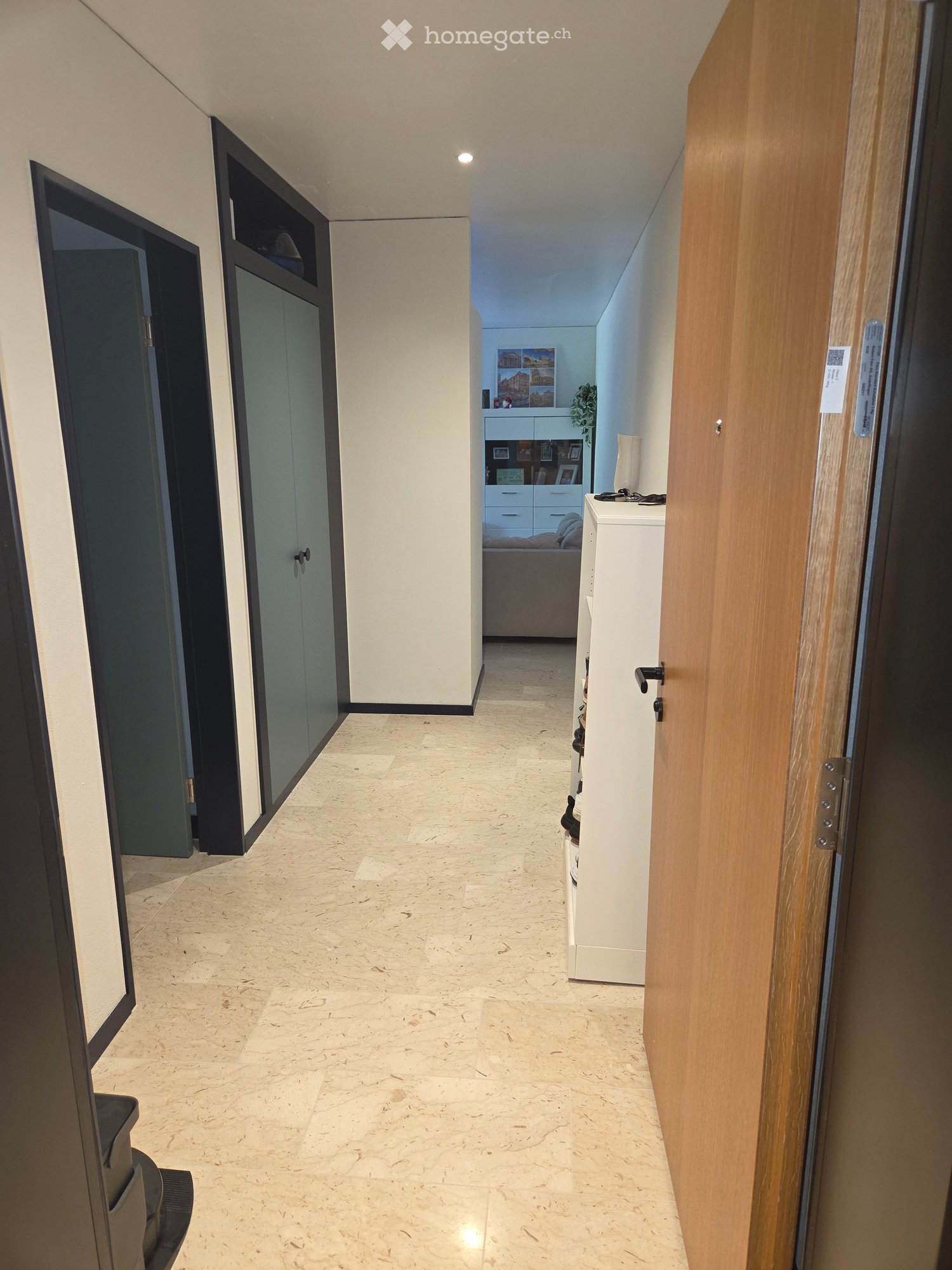 Modern hallway with marble flooring, white walls, and several doors leading to different rooms. A small table with a lamp and a plant is visible in the background.