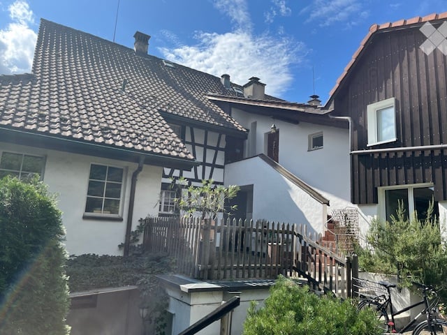 A multi-story residential building with a tiled roof, white walls, and wooden accents. The structure includes a staircase leading to an upper level, surrounded by greenery and a wooden fence.