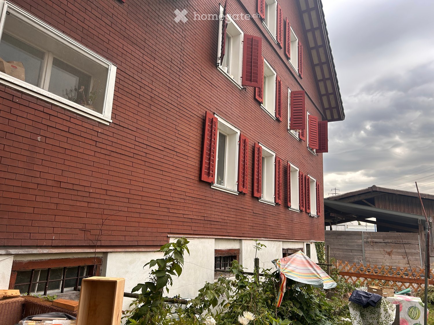 Two-story house with red brick exterior, white-framed windows, and red shutters. Visible garden area with plants, a wooden crate, and a small umbrella.