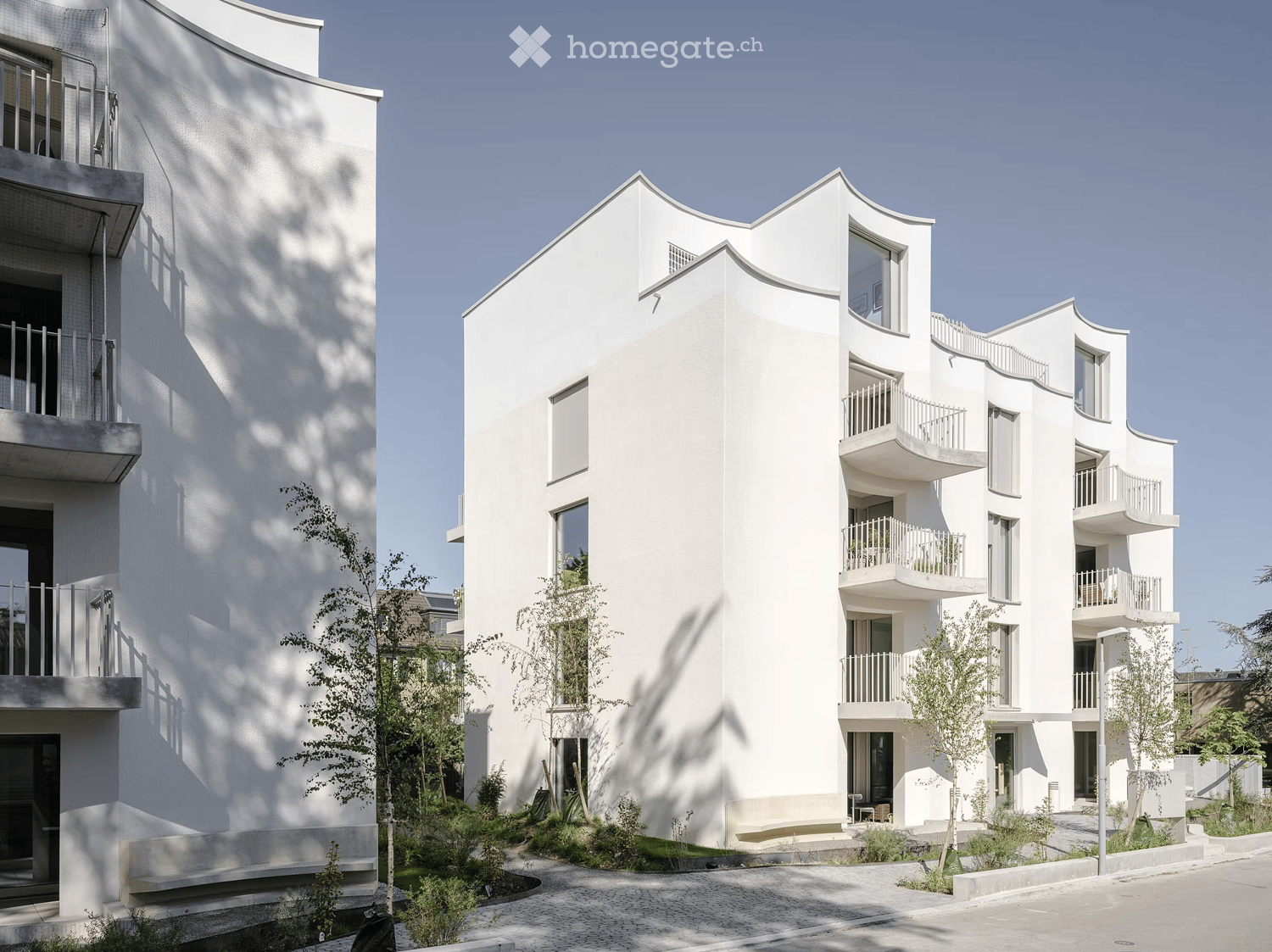 Modern white apartment building with multiple balconies and contemporary design, surrounded by small trees and greenery.