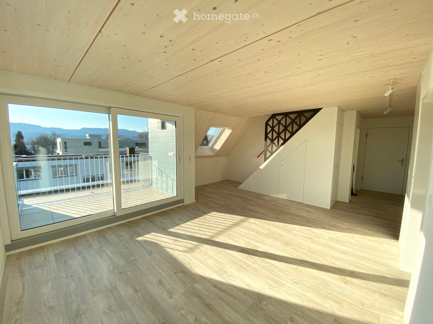 Bright, empty attic room with wooden floors and exposed wooden beams. Features large sliding glass doors leading to a balcony, a staircase leading to another level, and a skylight window.