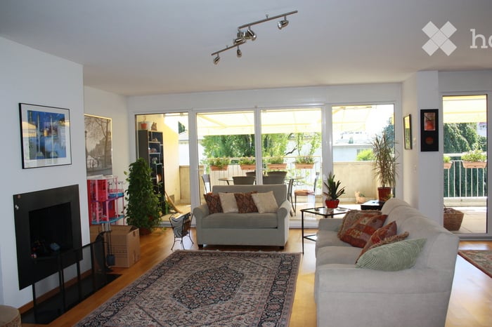 Bright living room with large sliding glass doors leading to a balcony, fireplace, two sofas, coffee table, bookshelf, and decorative plants.