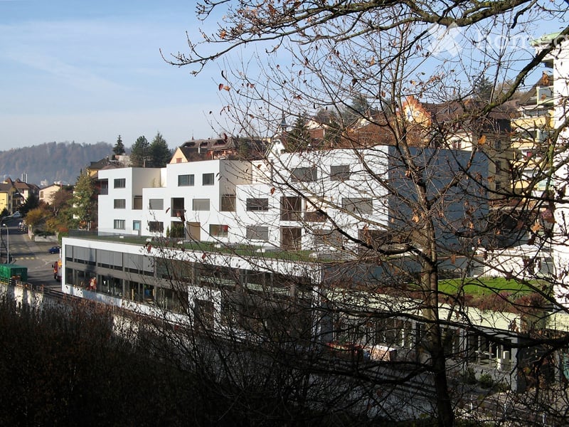 Modern multi-story building with white facade and large windows, surrounded by trees and other structures in an urban setting.