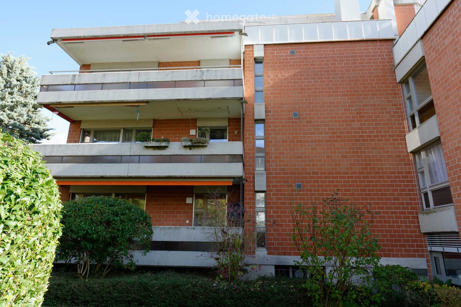 A multi-story brick building with balconies, large windows, and greenery around the base.