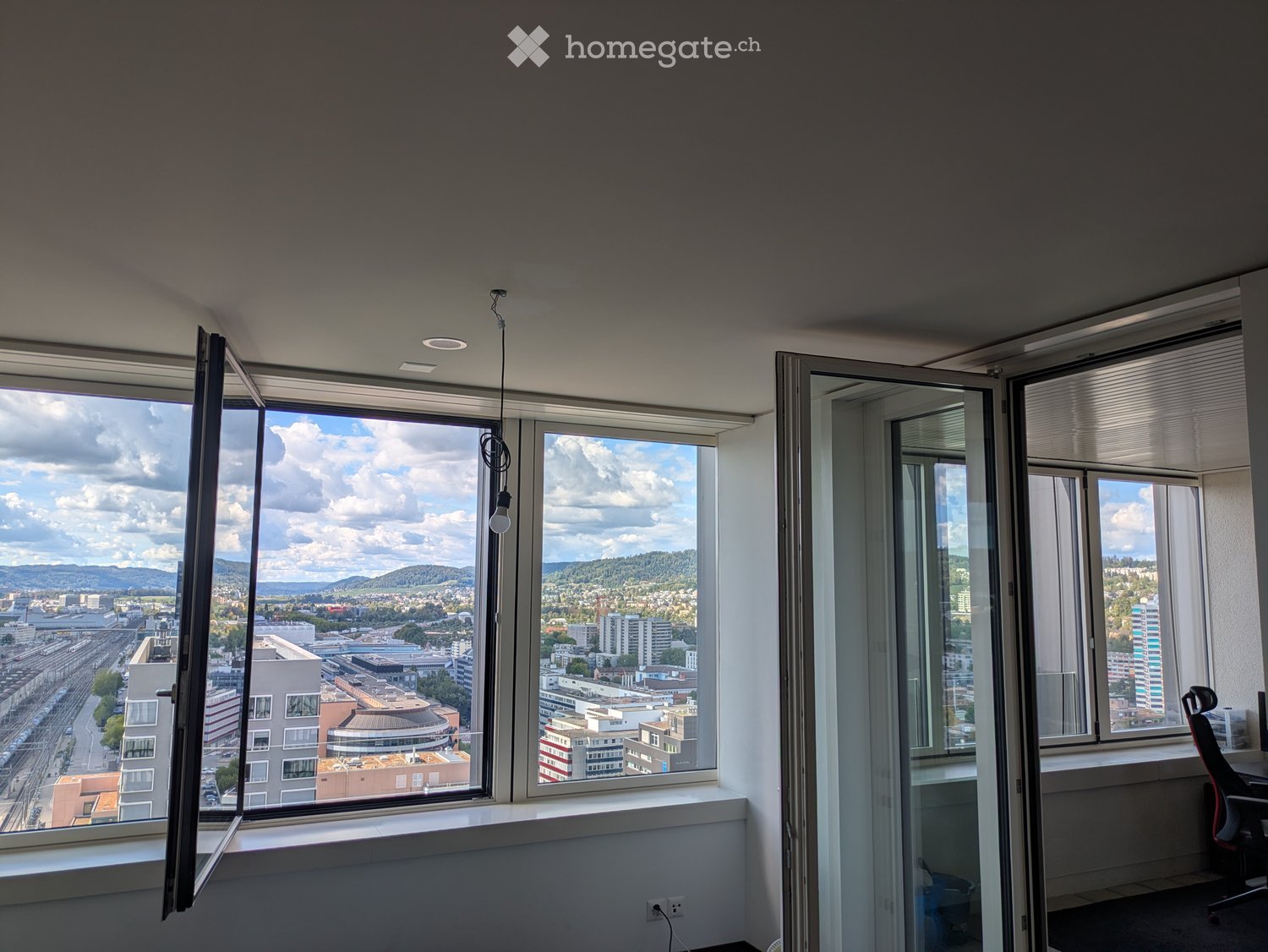 Empty room with large windows offering a cityscape view, white walls, and a hanging light fixture.