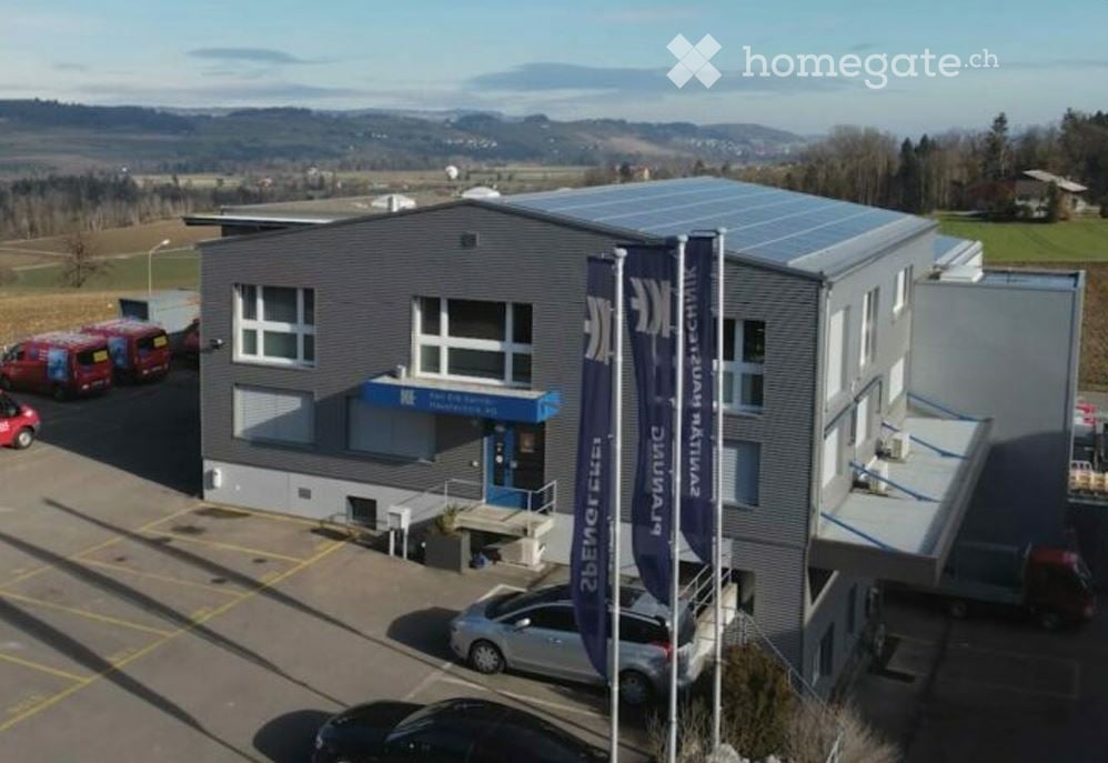 Modern industrial-style building with solar panels on the roof, multiple windows, and a blue entrance sign. Several vehicles are parked in the lot.