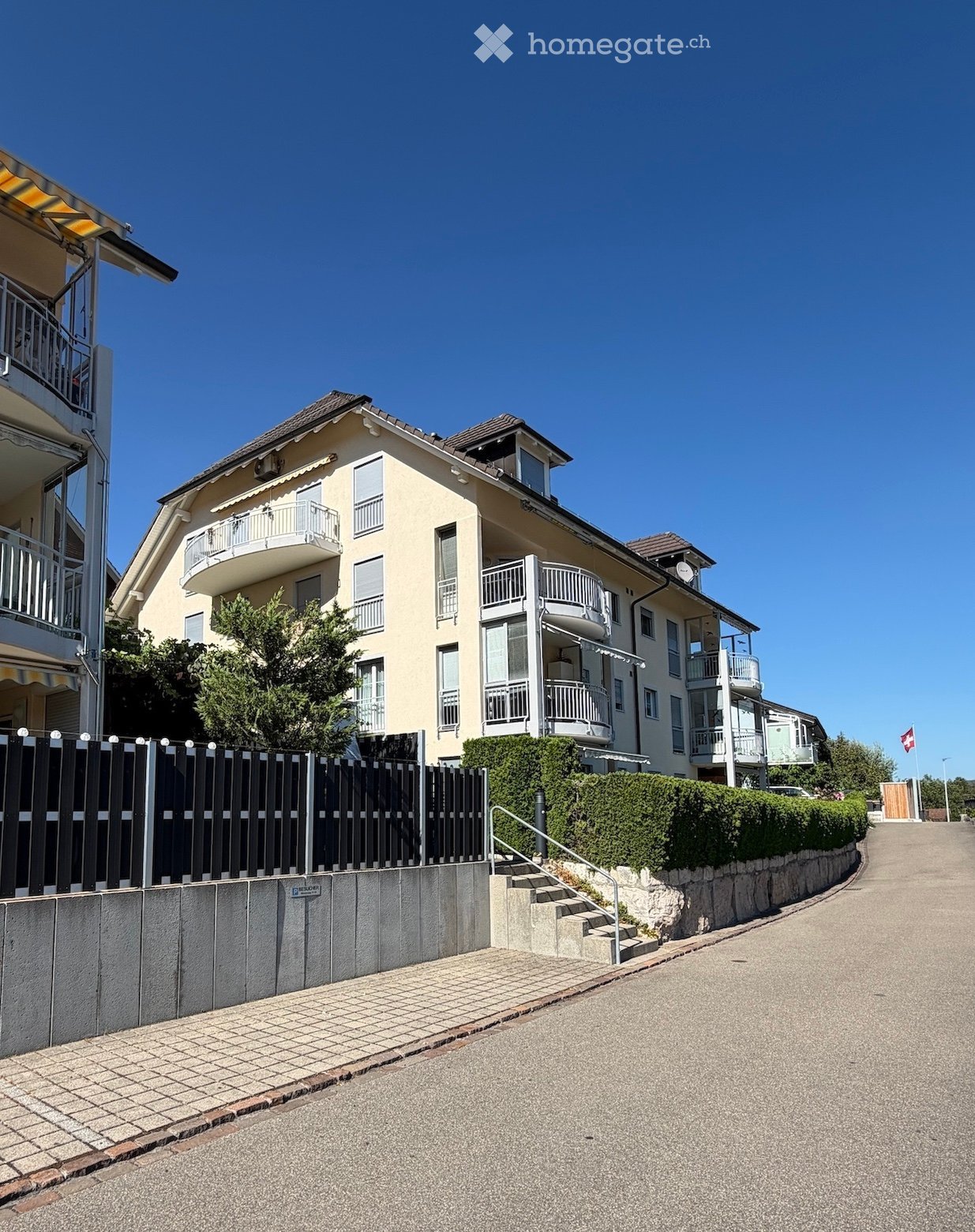 A two-story residential house with a light yellow facade, multiple balconies, and a well-maintained garden area with a low hedge and a black fence.