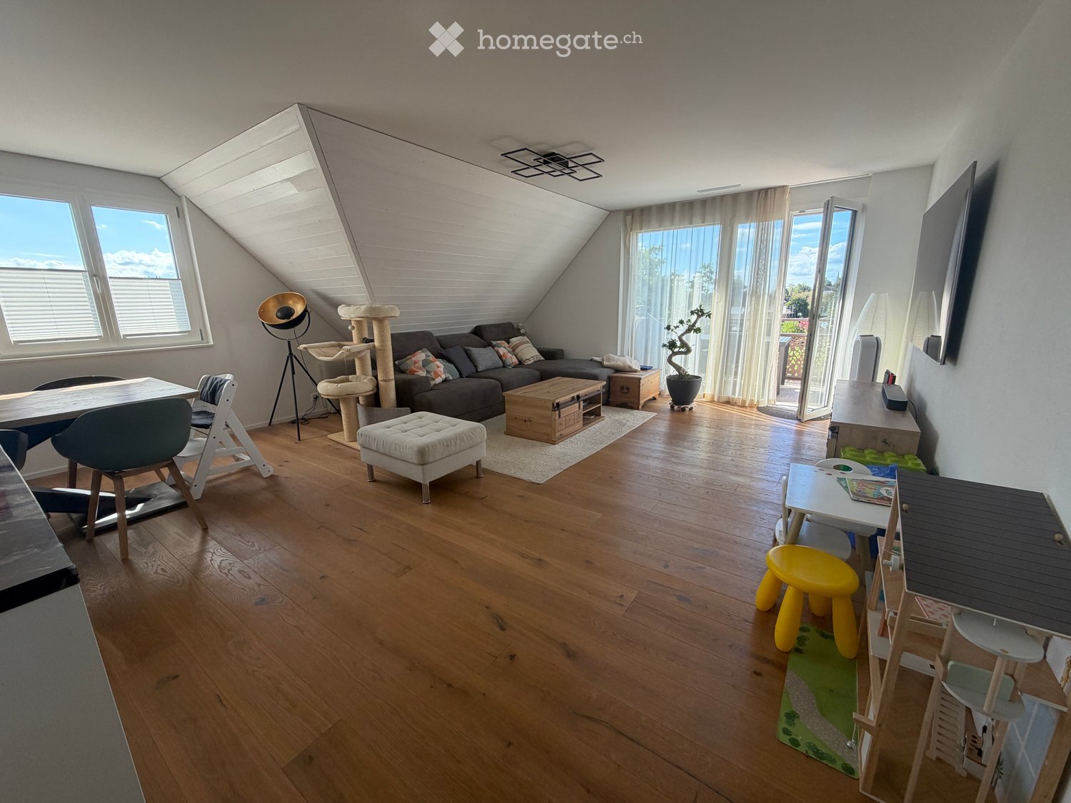 Bright, open living space with wooden flooring, large windows, and skylight. Features a sofa, ottoman, coffee table, cat tree, dining table with chairs, and a small play area for children.