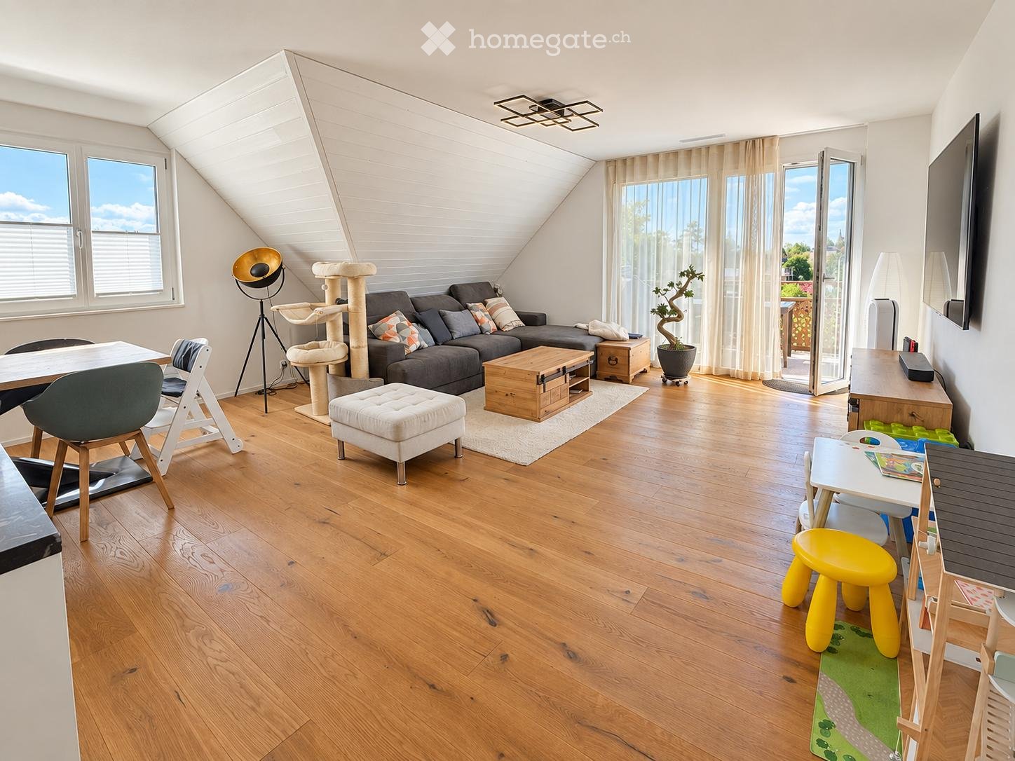 Bright living room with wooden floors, large windows, and skylights. Features a grey sectional sofa, coffee table, ottoman, cat tree, dining table with chairs, and a small play area with a yellow stool and green play mat.