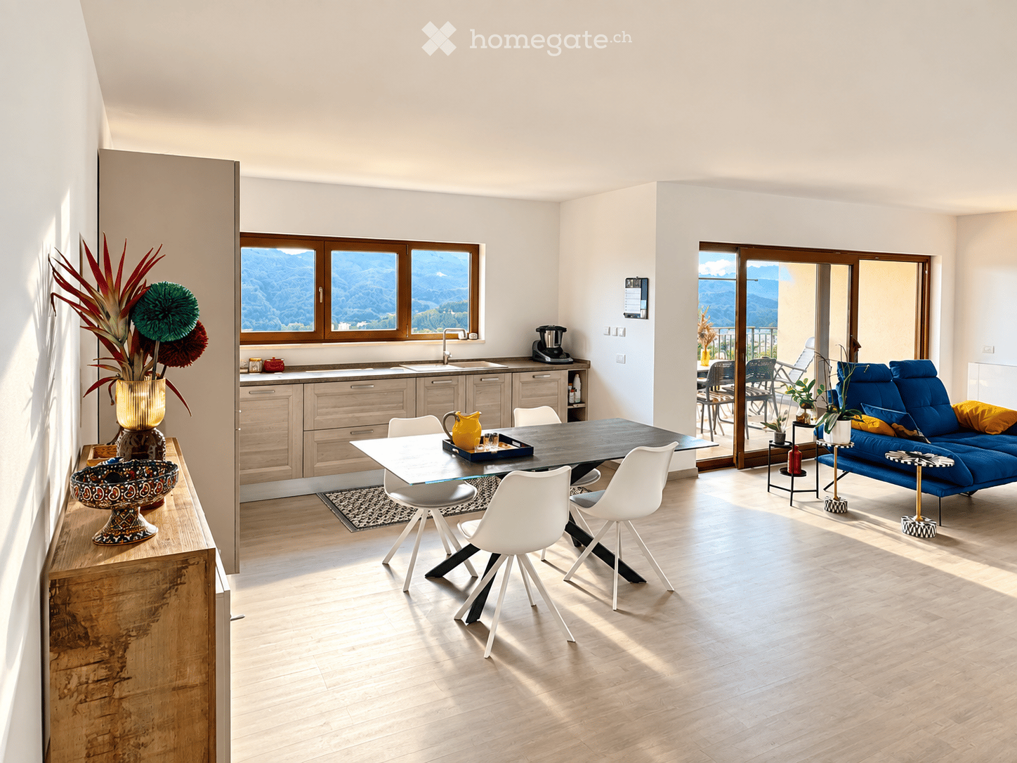 Open-plan living and dining area with modern furniture, large windows offering a scenic mountain view, and a balcony accessible through glass doors.