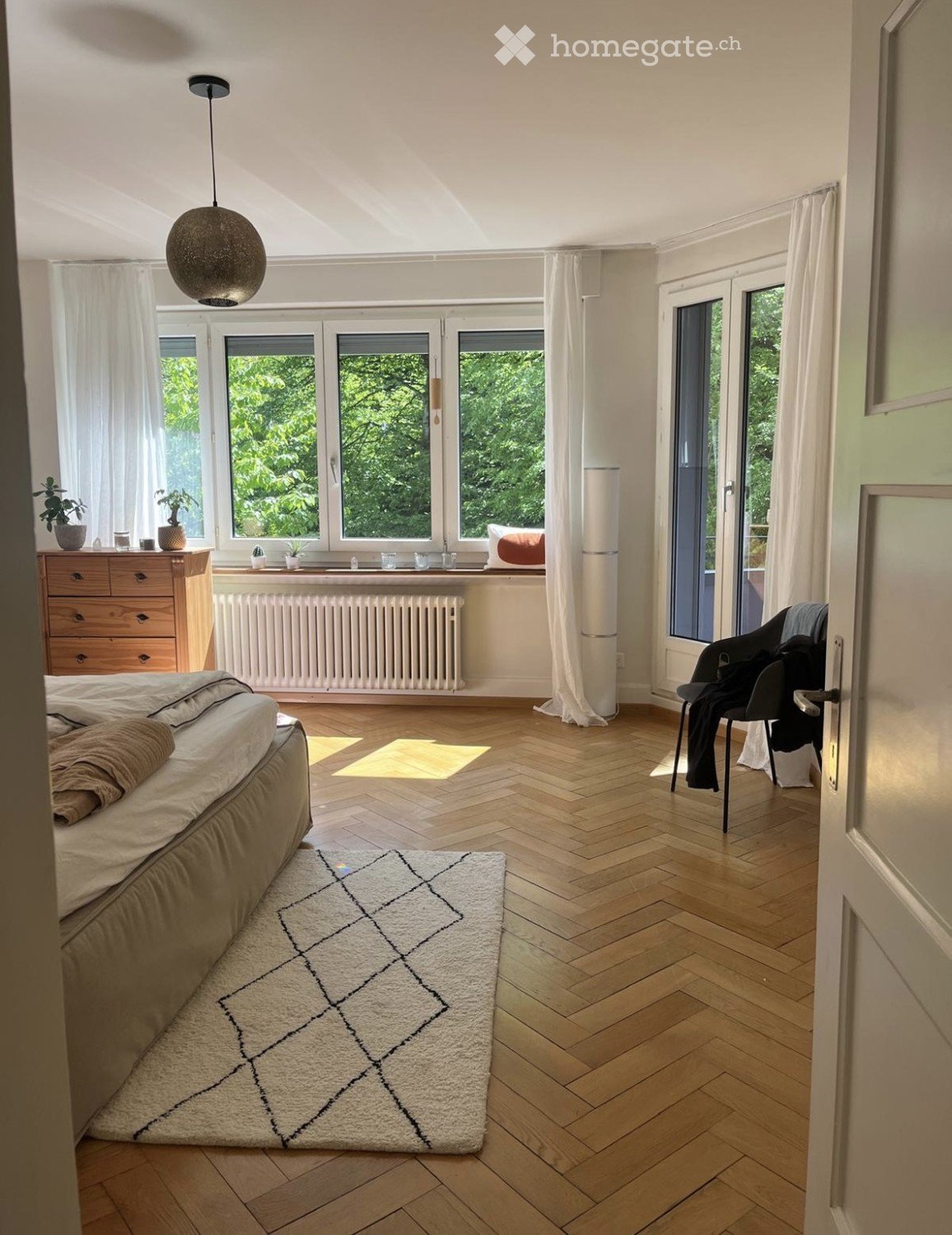 Bright bedroom with wooden herringbone flooring, large windows with white curtains, a bed with beige bedding, a patterned rug, a wooden dresser with potted plants, a chair with folded clothes, and a modern pendant light fixture.