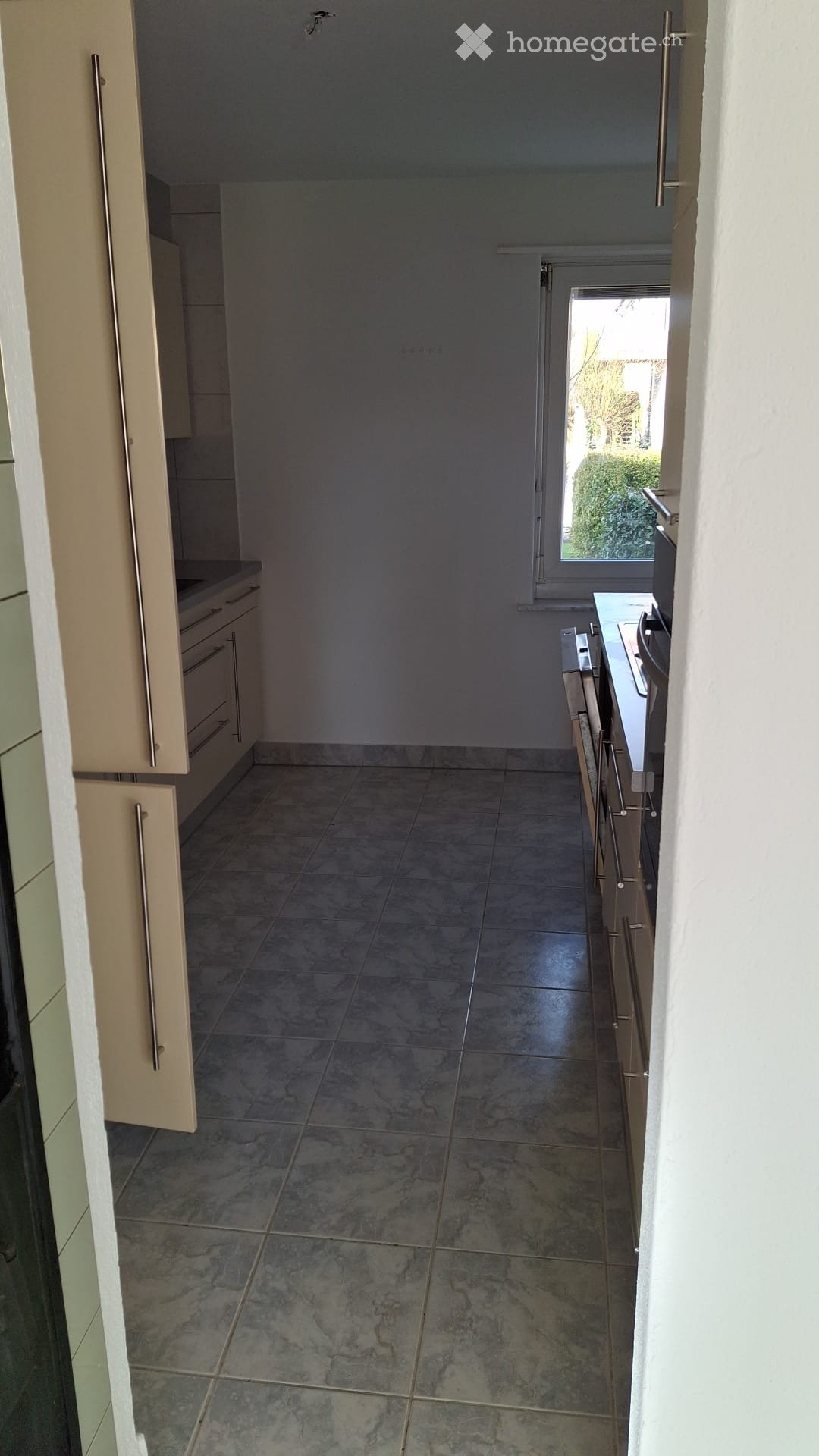 Empty kitchen with light-colored cabinets, tiled floor, and a window overlooking a garden area.