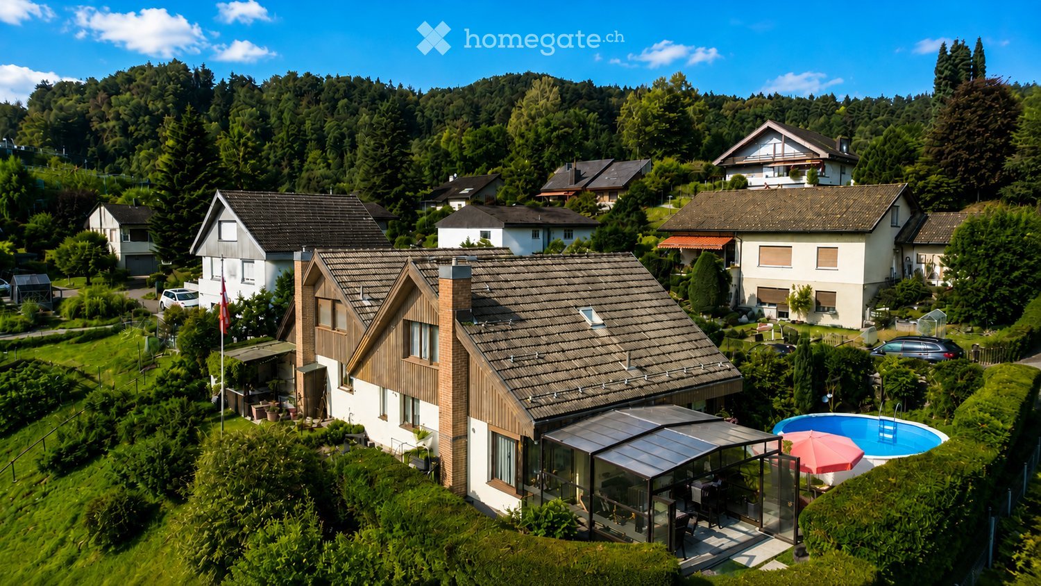 Aerial view of a modern house with a tiled roof, surrounded by greenery and neighboring houses. Features include a swimming pool, patio area, and outdoor furniture.
