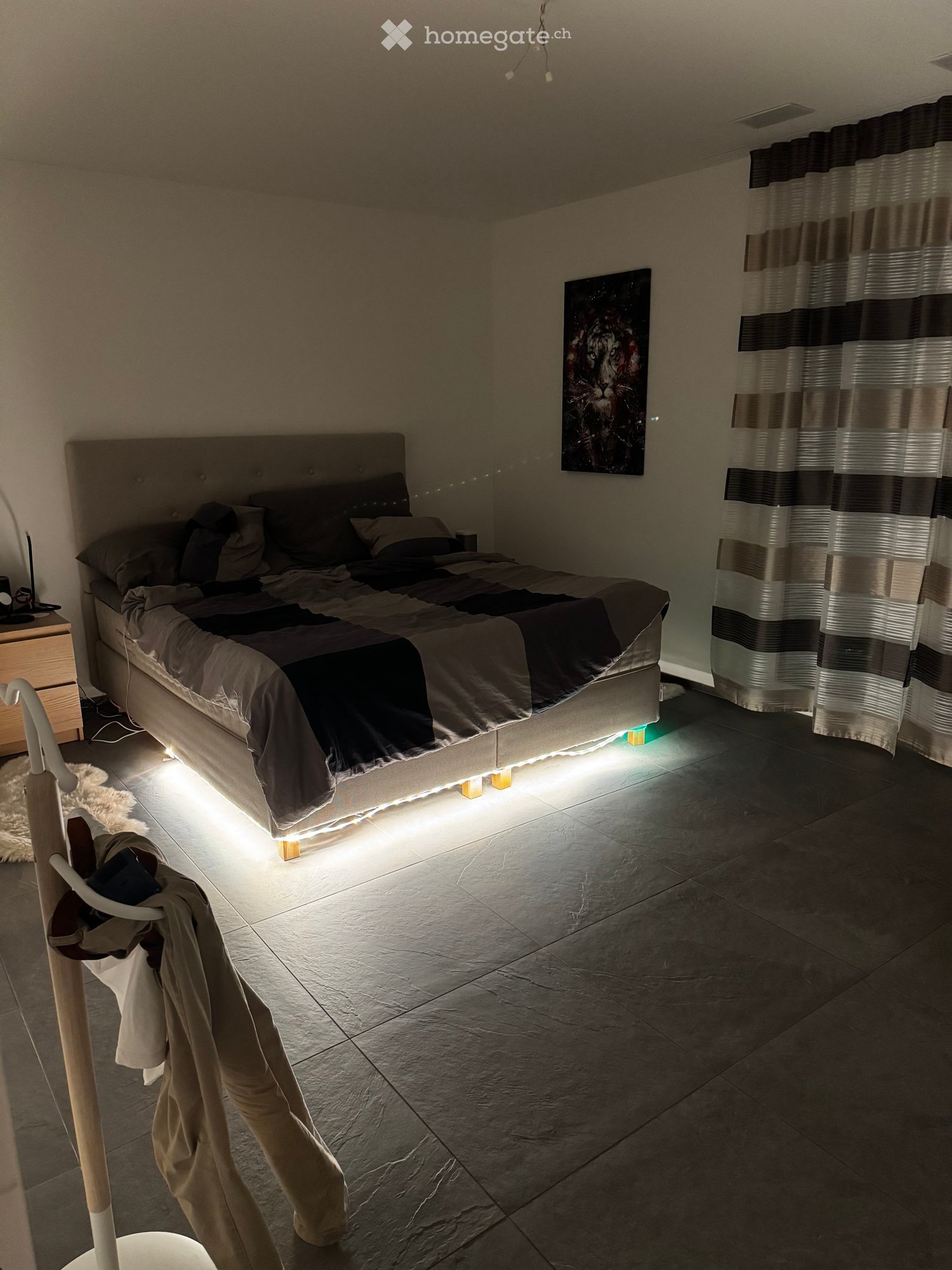 Modern bedroom with a large bed, nightstand, painting, and a curtained window. The room features tiled flooring and ambient lighting under the bed.