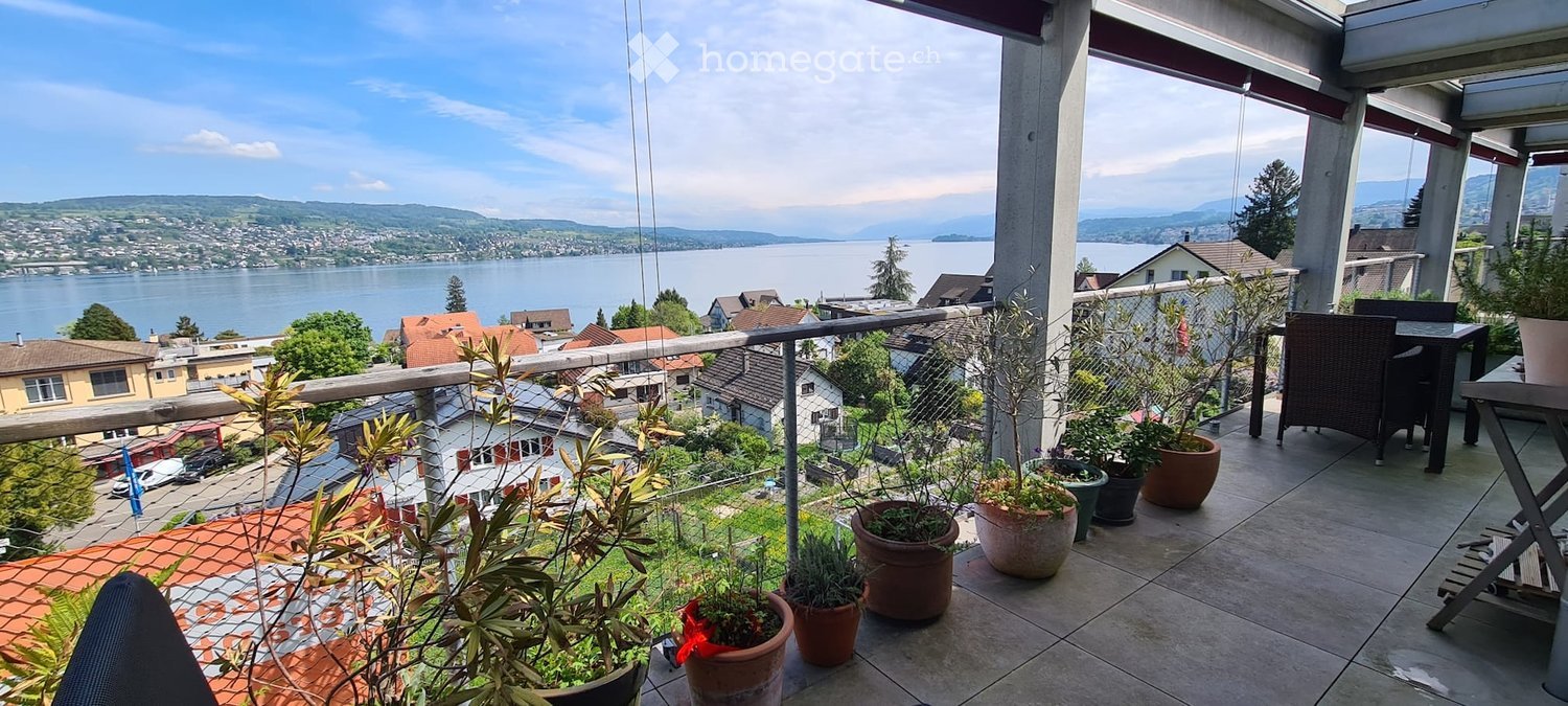 Balcony with a scenic view of a lake and surrounding town, featuring a table, chairs, and several potted plants.